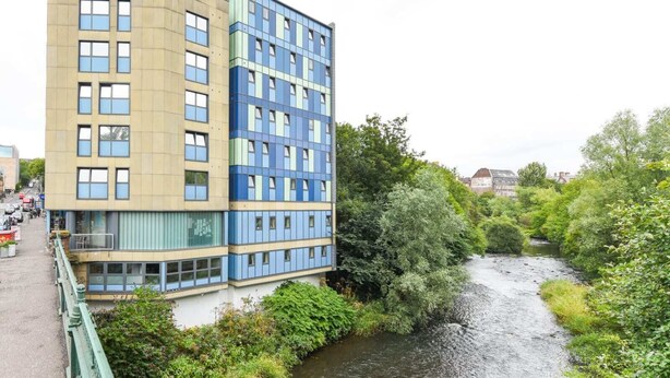 A modern building with a mix of beige and blue facades, a river in the foreground, and lush greenery. - Student Only Gibson Street (Glasgow)
