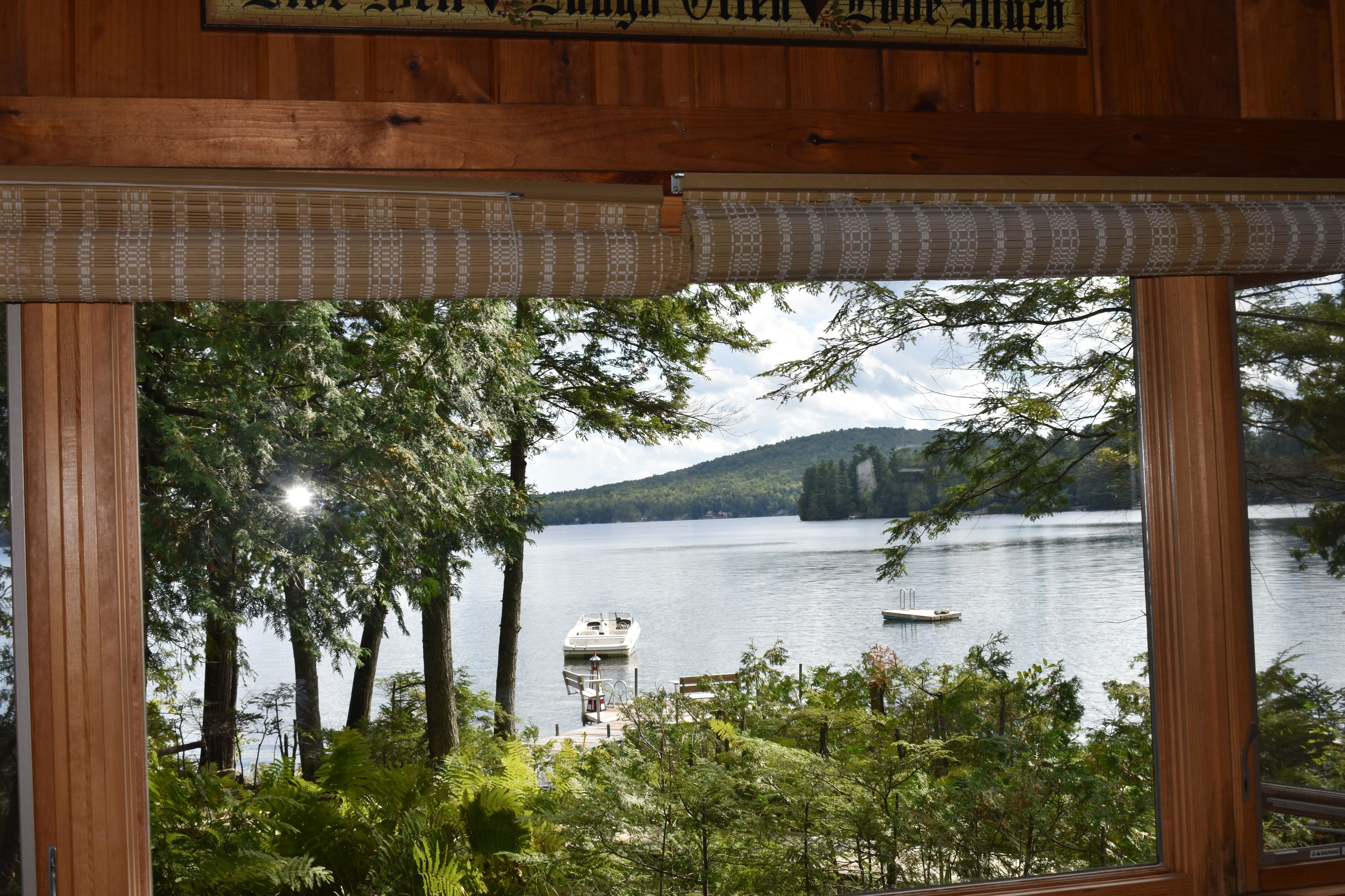 A view through a window with a wooden frame, overlooking a lake with a boat, trees, and a distant hill.