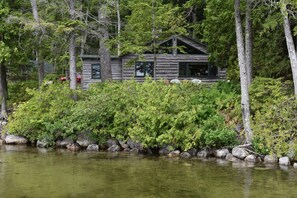 Une cabane aux murs en rondins de bois, entourée d’une forêt dense et bordée par un plan d’eau.