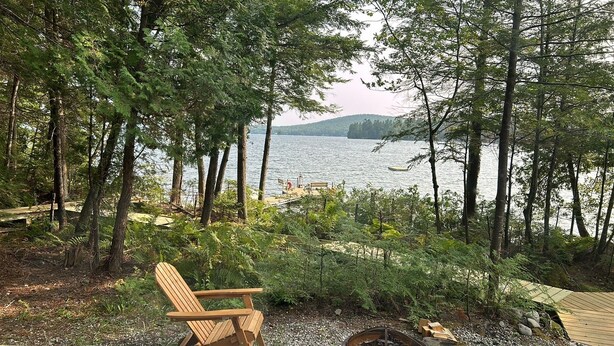 A wooden deck with a chair, surrounded by dense trees and overlooking a calm lake. - Welcoming 4-bedroom lake front cabin in charming Otis on Beech Hill Pond. (Otis)
