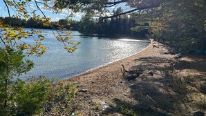 A lakeside view with a clear sky, surrounded by trees and a sandy shore. - Welcoming 4-bedroom lake front cabin in charming Otis on Beech Hill Pond. (Otis)