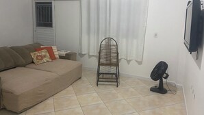 Ett vardagsrum med en beige soffa, en fåtölj i rotting, en vit gardin, en golvfläkt och en tv som är monterad på väggen.