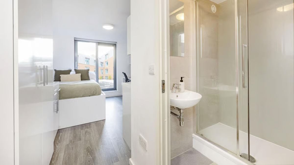 A modern bathroom with a glass shower enclosure, a white sink, and a bedside table with a lamp.