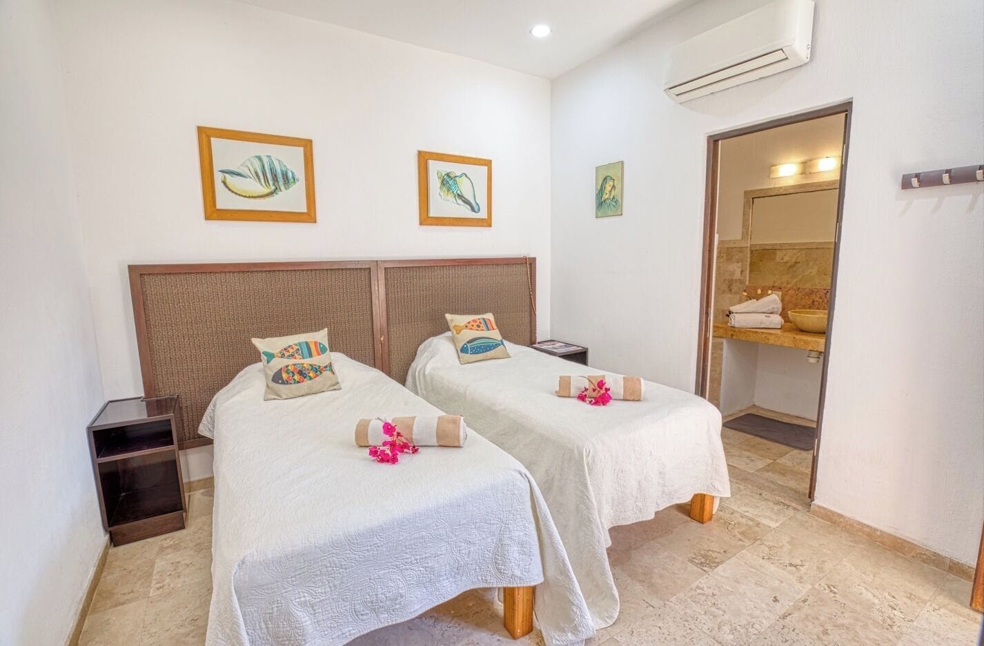 A hotel room with two beds, a small bedside table, an air conditioning unit, and a bathroom with a mirror and sink.