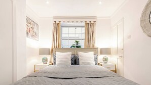Un dormitorio ordenado con una cama grande, dos mesitas de noche con lámparas, una ventana con cortinas y un adorno en la pared.