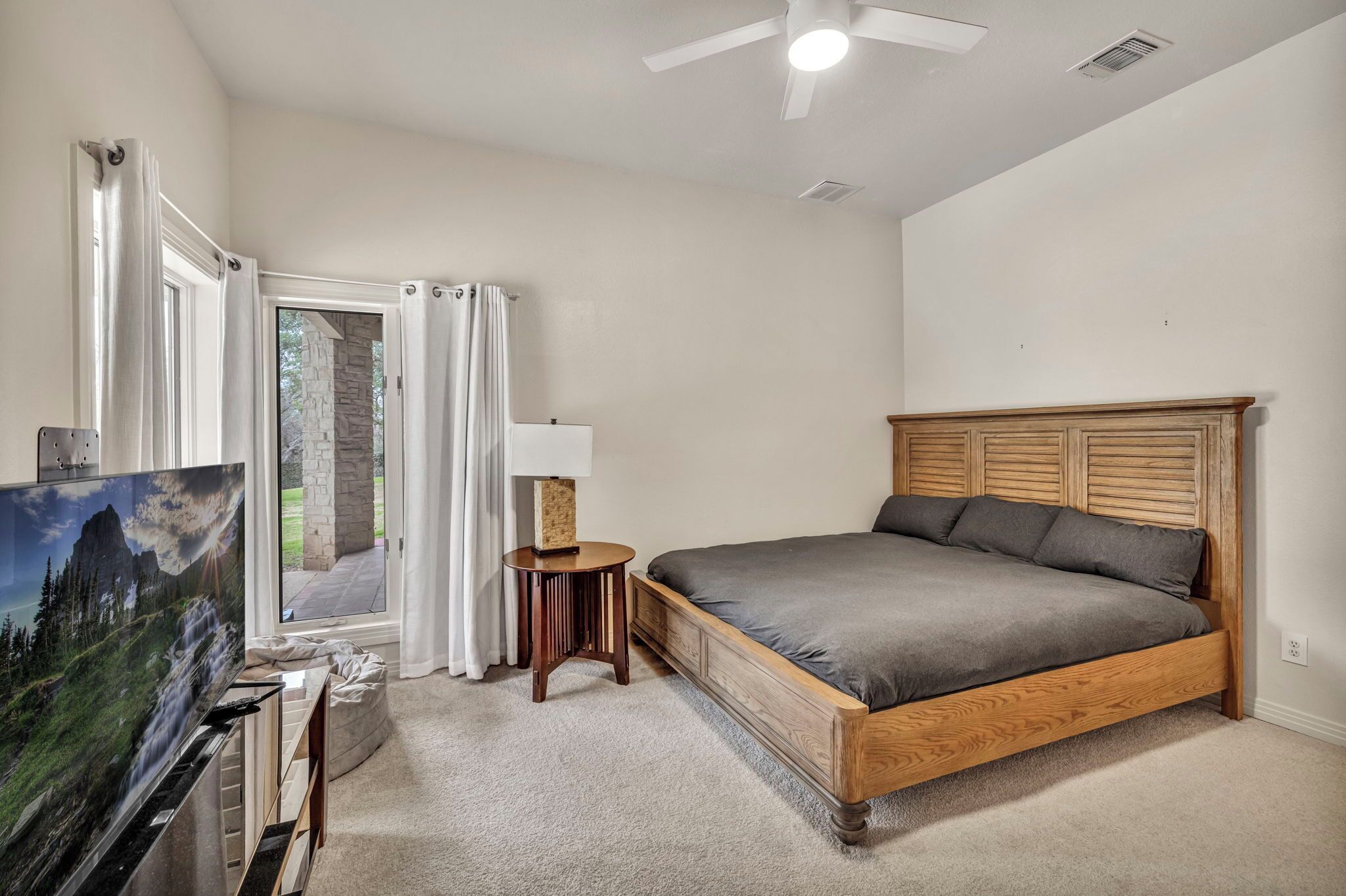 A bedroom with a wooden bed, a television, a small round table, and a view of a stone building through a glass door.