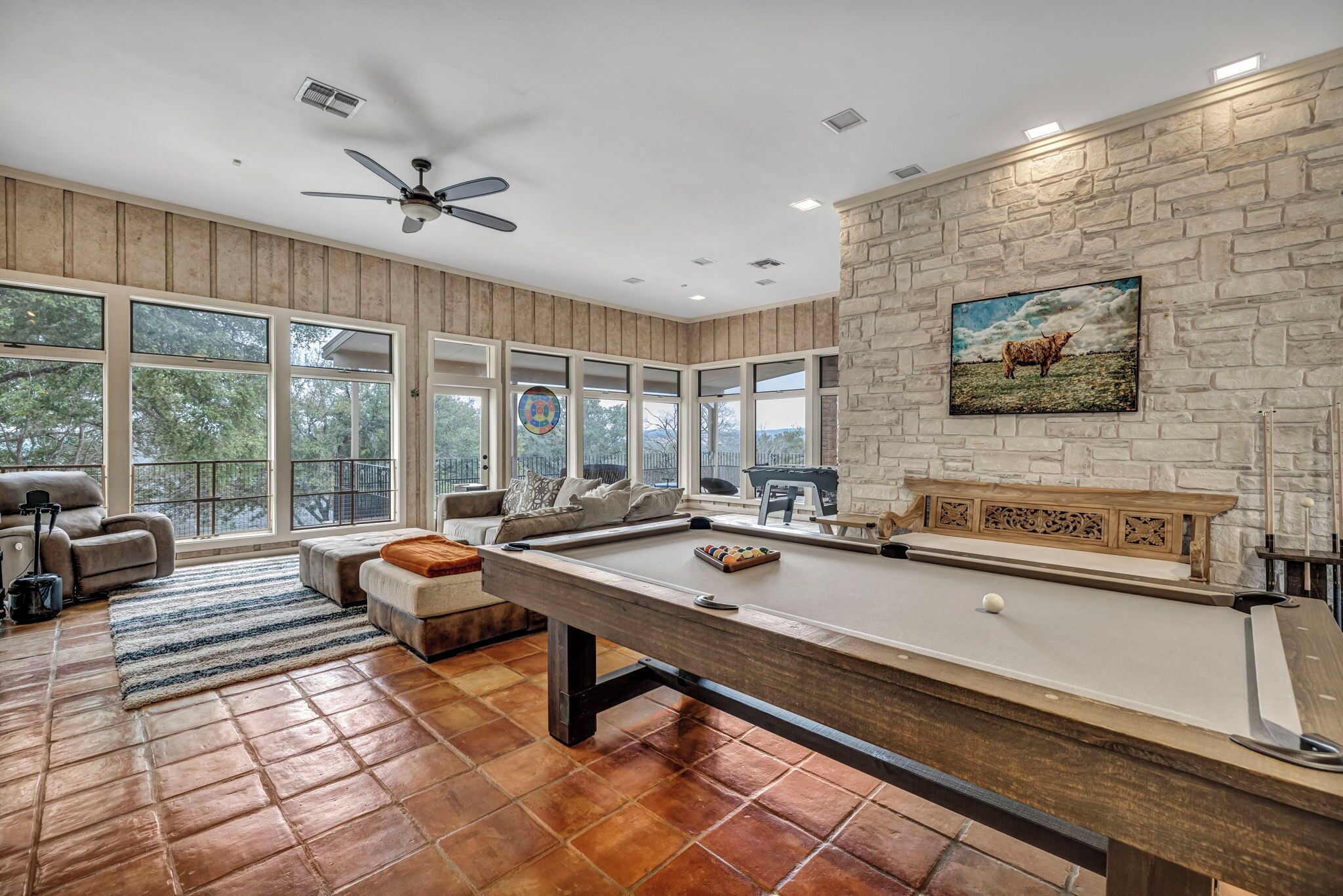 Une table de billard dans une pièce dotée de grandes fenêtres, d’un ventilateur de plafond et d’un mur en pierre orné d’un tableau.