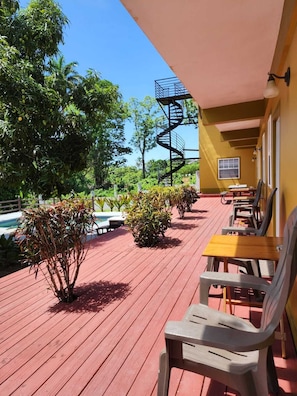 Une terrasse en bois avec des sièges d’extérieur, un escalier en colimaçon et une piscine en arrière-plan.