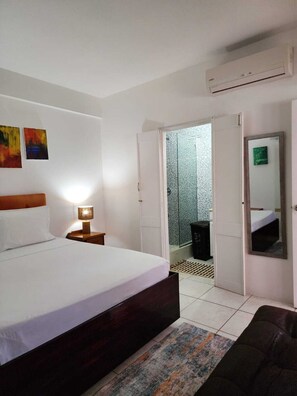 A hotel room with a bed, bedside lamp, a small table, a mirror, and an air conditioning unit.