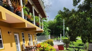 A wooden deck with outdoor seating, a yellow building with windows, and surrounding greenery. - Tropical Apartments Tobago: Bluebell Apt (Carnbee)