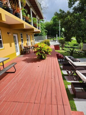 A wooden deck with outdoor seating, a yellow building with windows, and surrounding greenery.