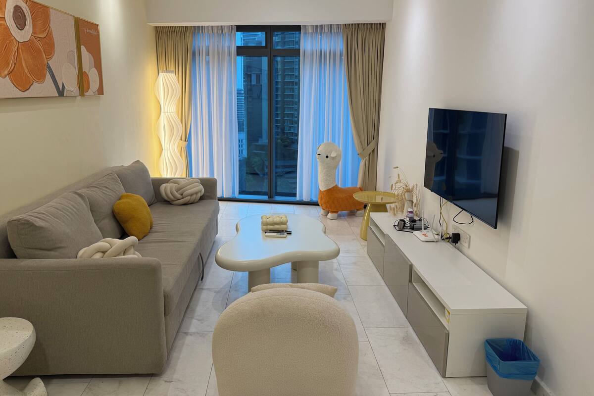 Una sala de estar moderna con un sofá, una mesa de centro, un televisor y un amplio ventanal con cortinas.