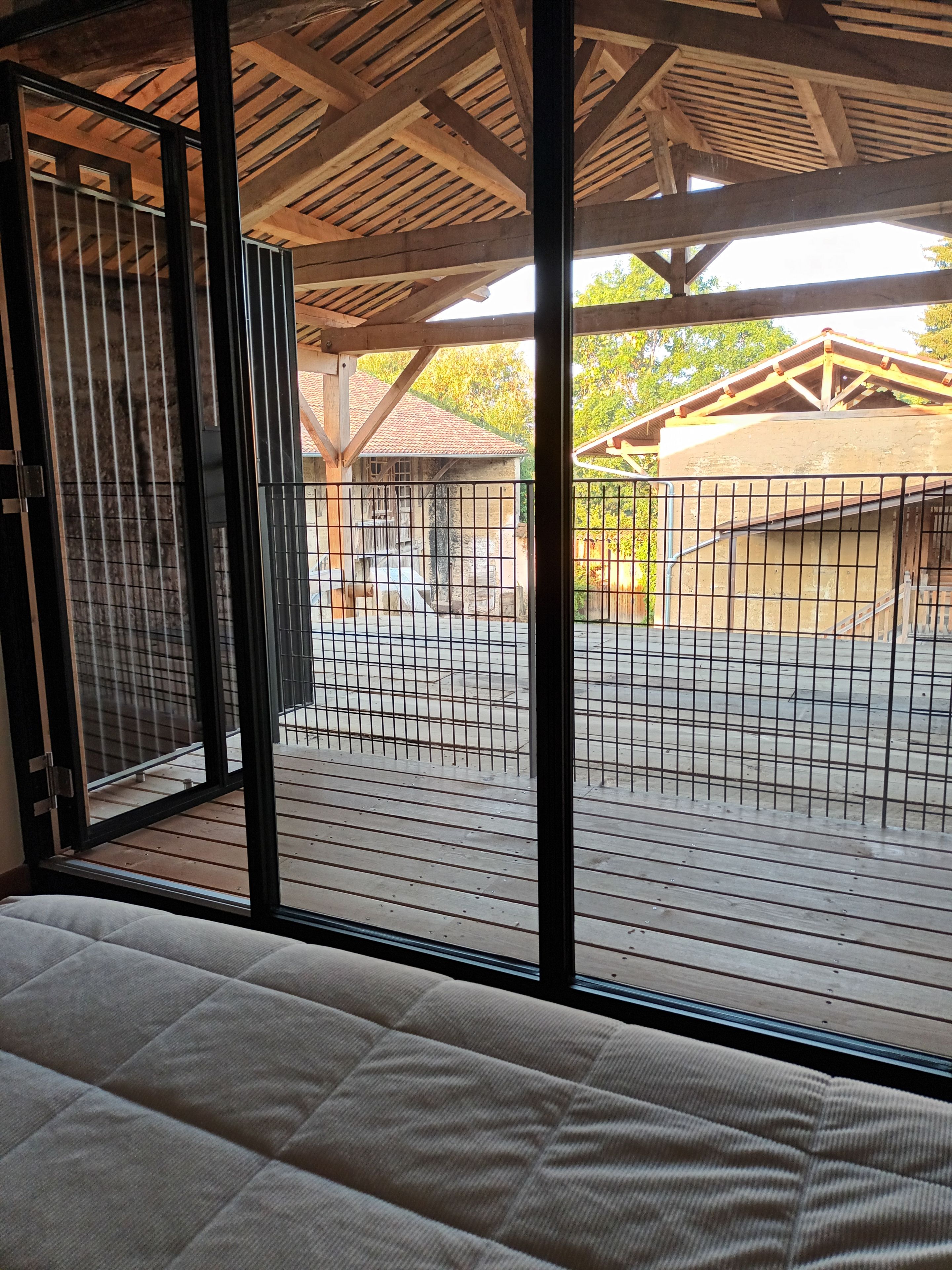 A room with a large window, wooden ceiling beams, and a view of an outdoor area with a fence and buildings.