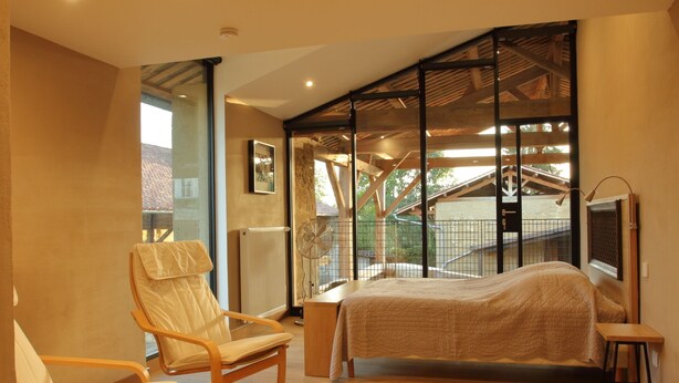 A bedroom with a bed, a chair, a radiator, and a window with a view of a building and trees. - La ferme de Berlioz (La Cote-Saint-Andre)