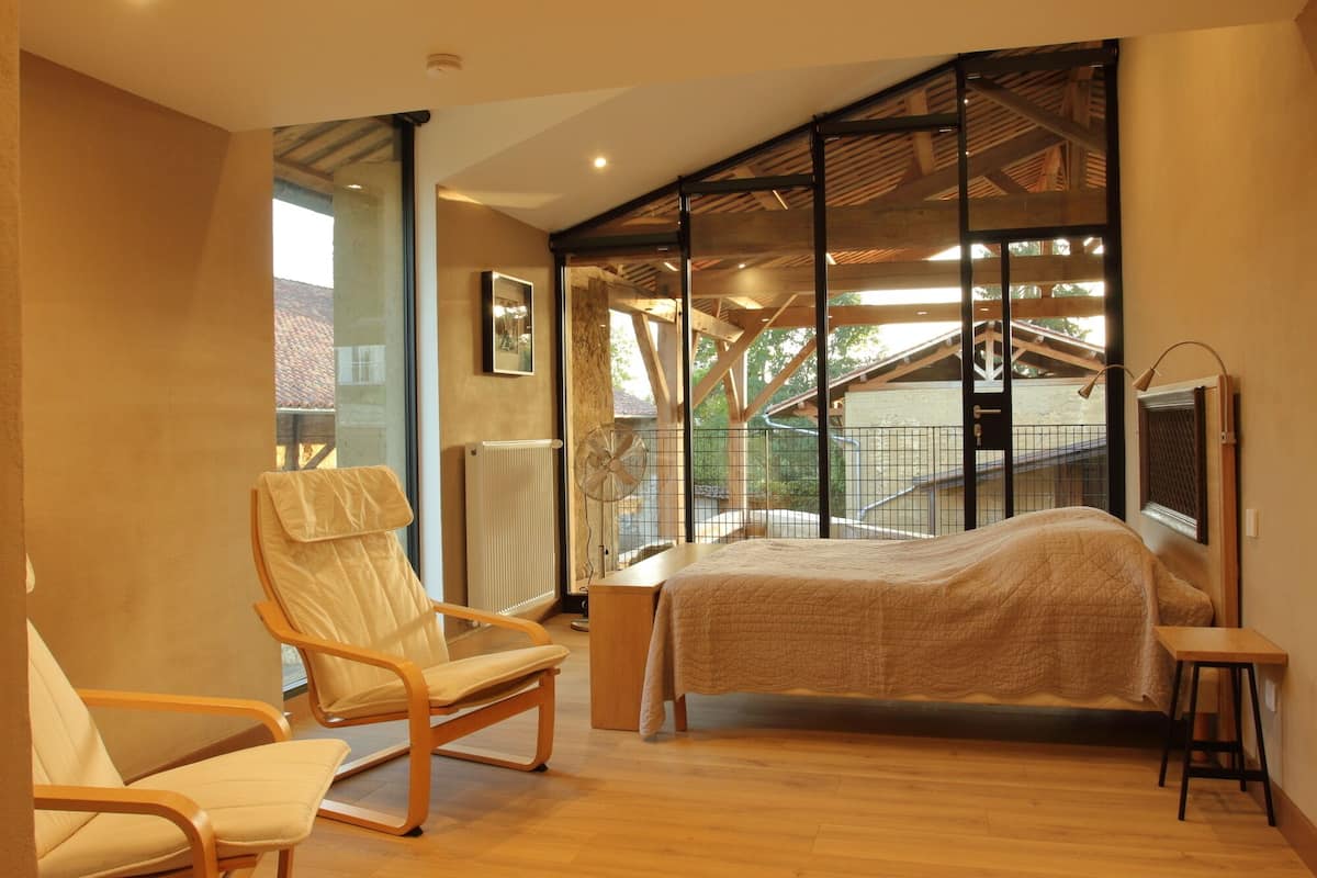 A bedroom with a bed, a chair, a radiator, and a window with a view of a building and trees.