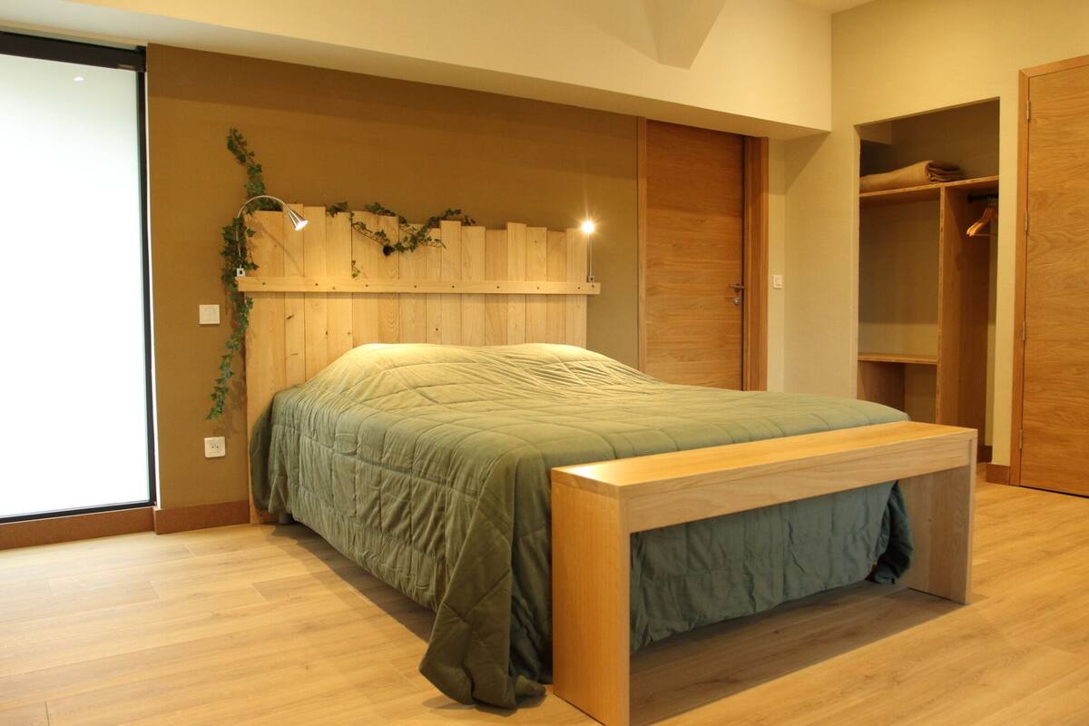 A bedroom with a wooden bed frame, a bench at the foot of the bed, a wardrobe, and a wooden wall with a plant.