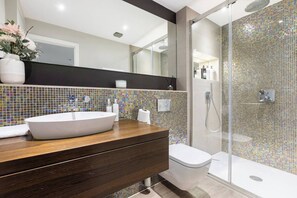 A modern bathroom with a large white sink, a wooden vanity, a glass shower enclosure, and a tiled backsplash. - Charming 4 Bed Townhouse Opposite Buckingham Palace (London)