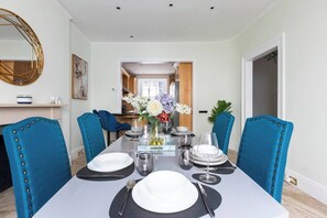 A dining room with a rectangular table set for six, blue upholstered chairs, and a vase with flowers. - Charming 4 Bed Townhouse Opposite Buckingham Palace (London)