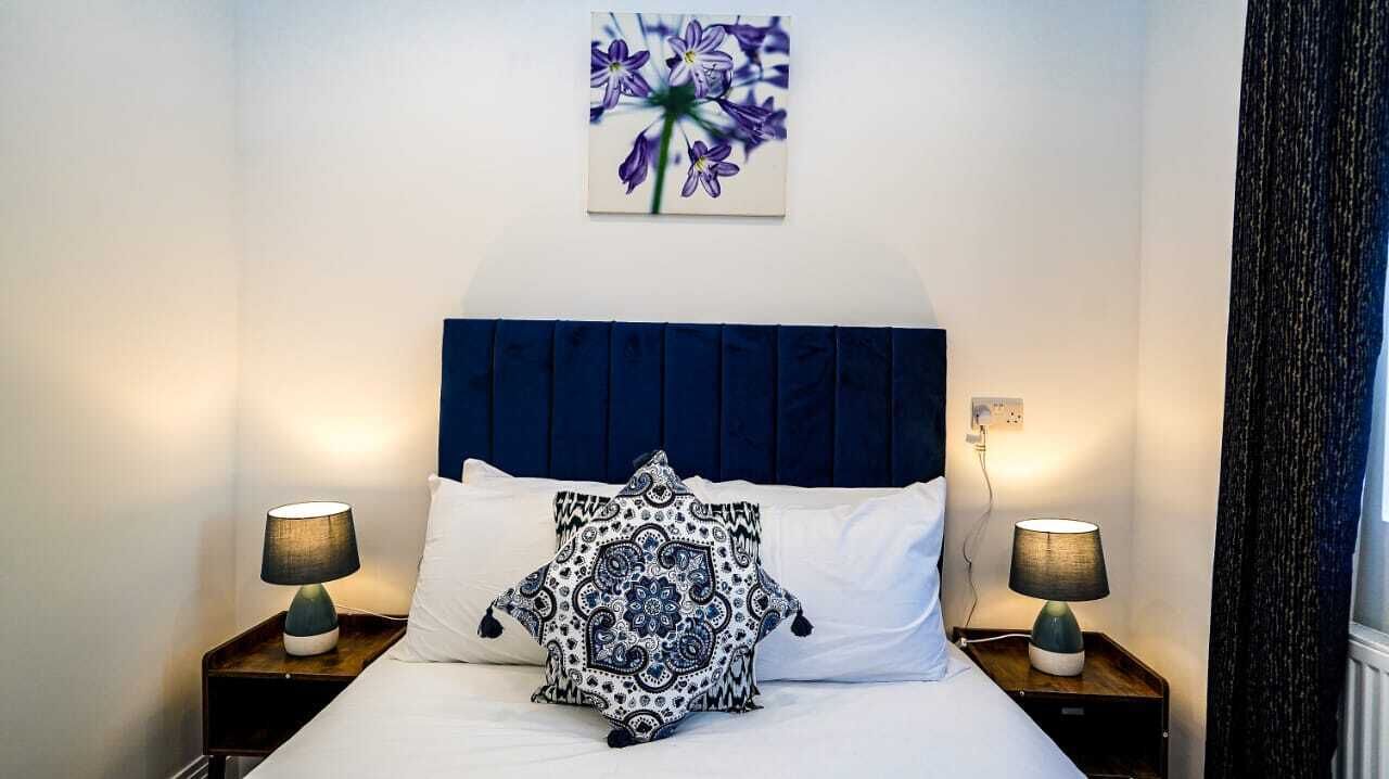 Un lit bien fait, agrémenté d’un coussin à motifs, de deux lampes de chevet et d’un tableau représentant des fleurs accroché au mur.