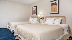 Habitación de hotel con dos camas, cada una con cabecera, ropa de cama blanca y una lámpara en la mesita de noche.