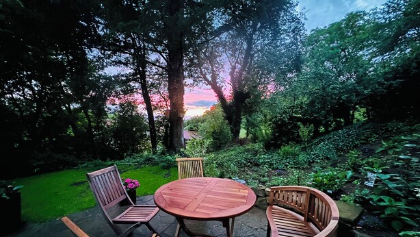 Un espacio al aire libre con una mesa redonda de madera y cuatro sillas, rodeado de abundante vegetación y árboles.