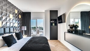 A modern hotel room with a large bed, a TV mounted on the wall, and a balcony with outdoor seating.