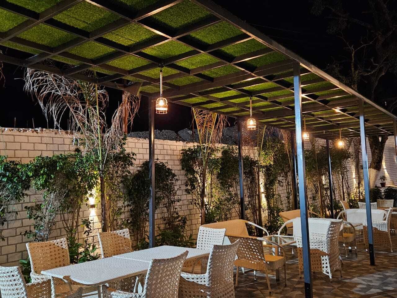 An outdoor patio area with tables and chairs, covered by a pergola with a green roof, and walls adorned with climbing plants.