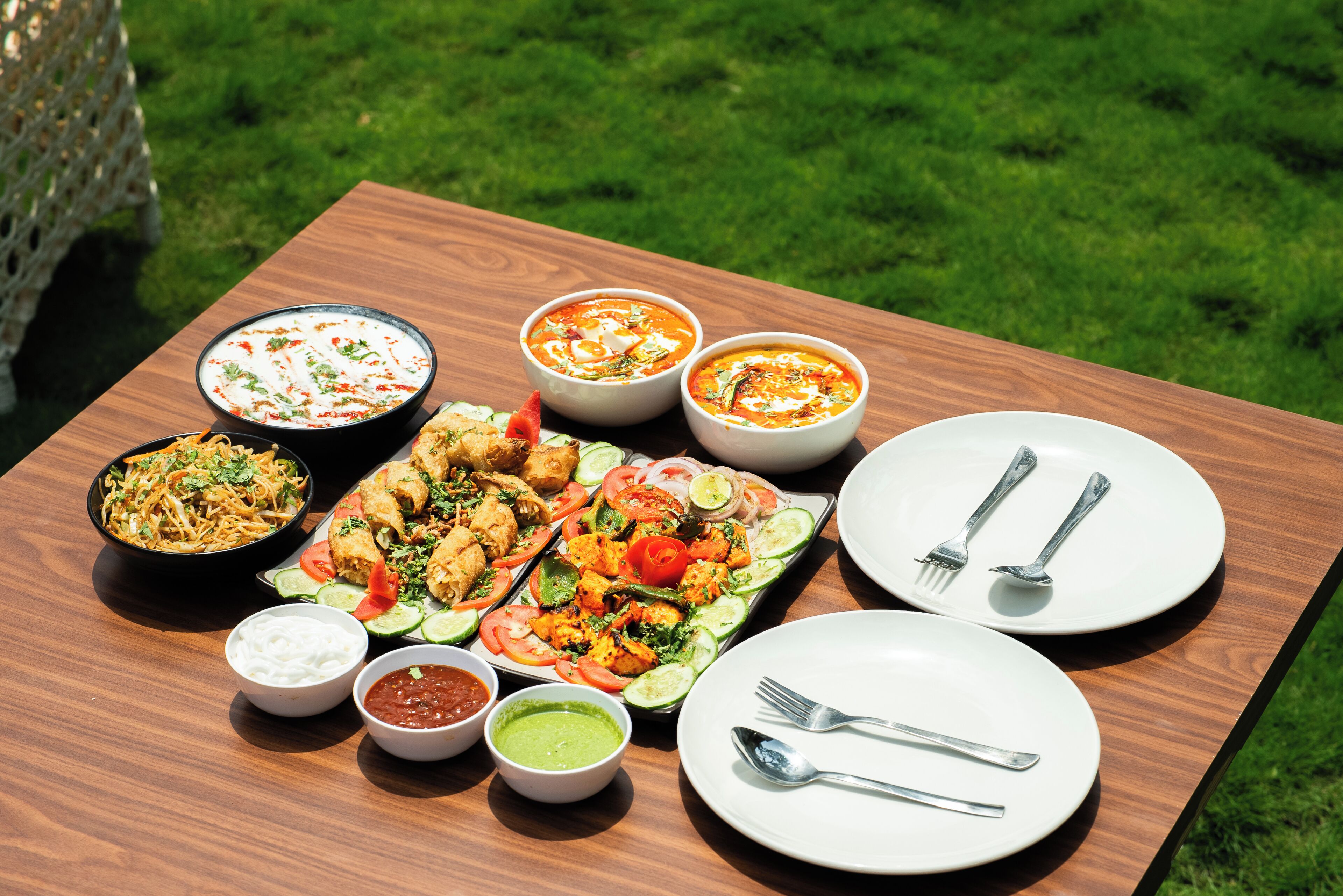 A wooden table with various dishes, including a salad, grilled chicken, and bowls of soup, set outdoors on a grassy area.