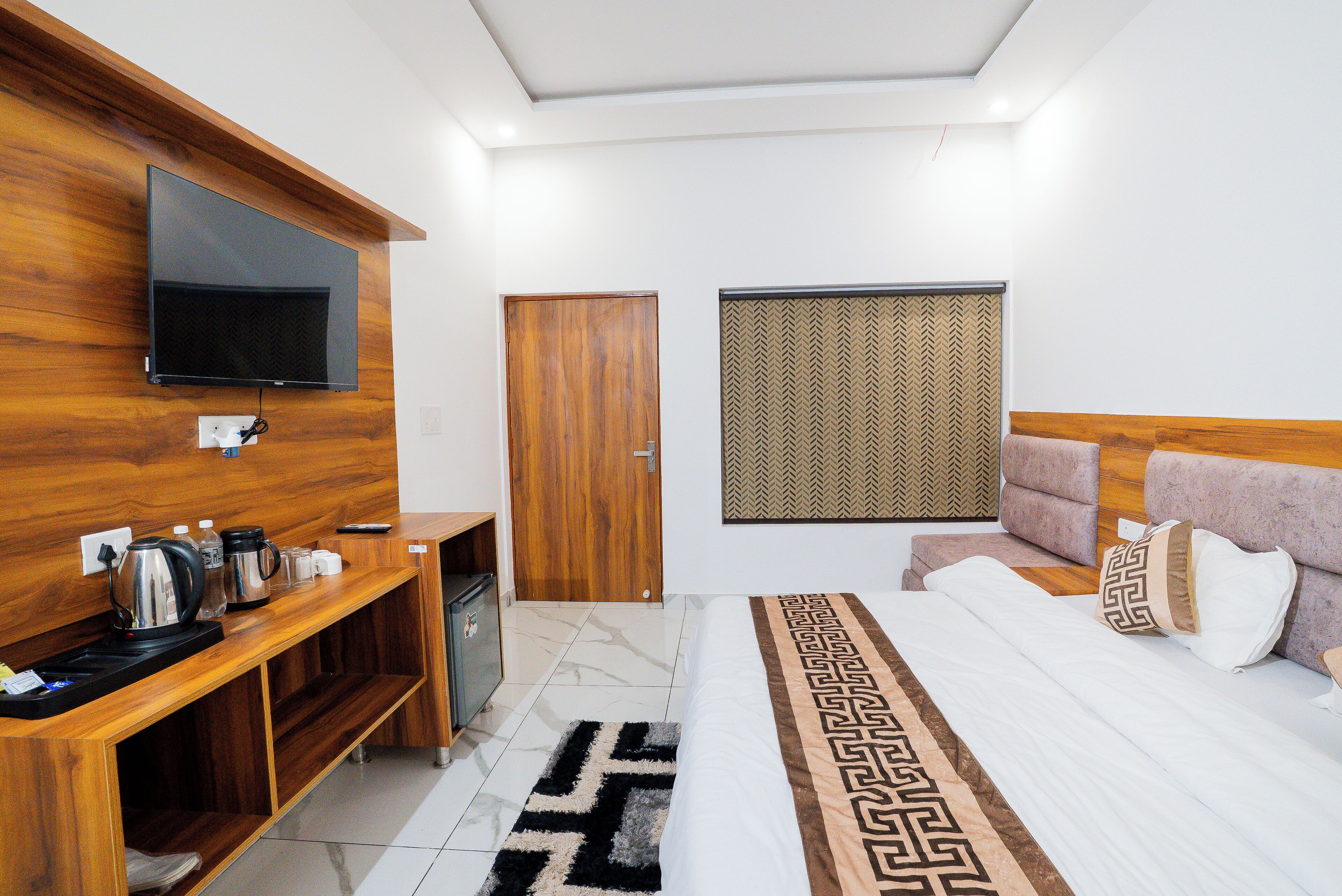 A modern hotel room with a flat-screen TV mounted on a wooden wall, a bed with a patterned runner, a wooden headboard, and a small kitchenette area.