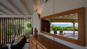 Una habitación con un tocador de madera, un espejo grande que refleja una vista al exterior, una silla de mimbre y una pequeña estatua.