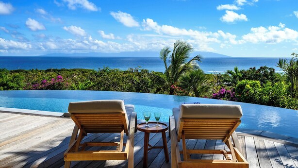 Una terraza de madera con dos sillones de descanso y una mesita con vasos, con vista a una piscina y un paisaje tropical que incluye el océano y montañas.