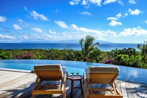 A wooden deck with two lounge chairs and a small table with glasses, overlooking a pool and a tropical landscape with a view of the ocean and mountains.
