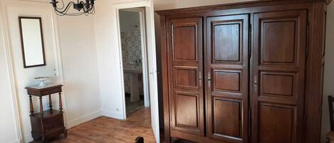 A room with wooden flooring, a large wooden wardrobe, a small wooden side table, and a mirror on the wall.