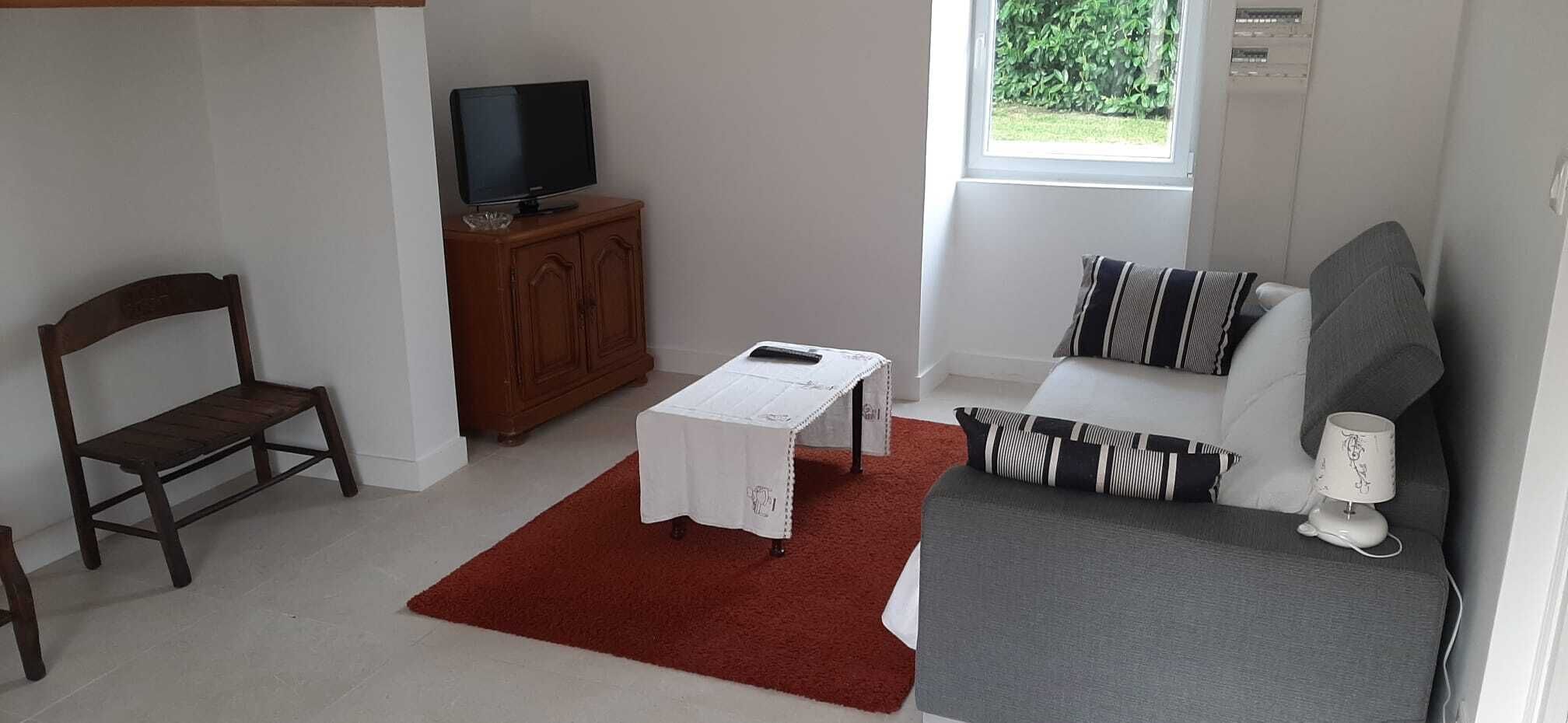 A small, well-lit room with a wooden chair, a small table with a white tablecloth, a sofa with striped pillows, and a television on a wooden cabinet.