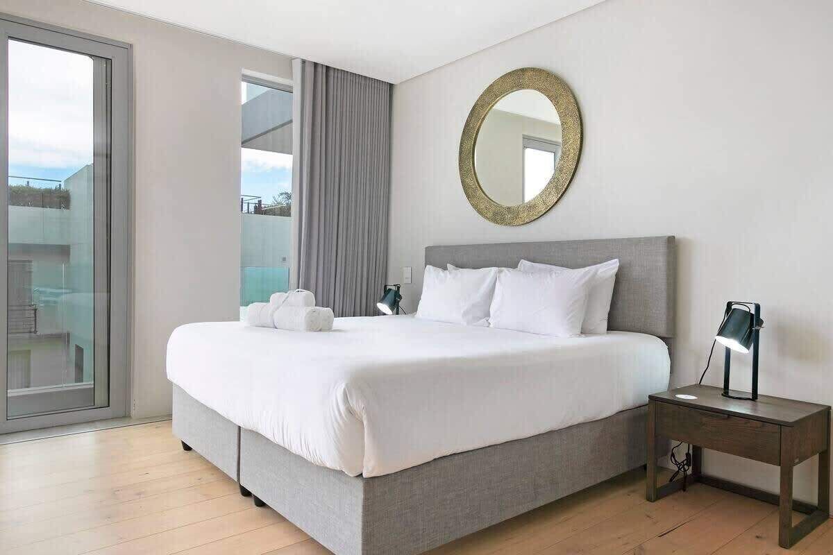 A modern hotel room with a large bed, a bedside table with a lamp, a round mirror on the wall, and a view through a large window.