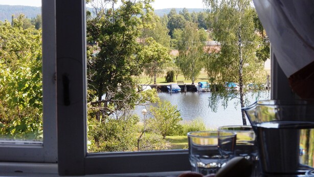 A view from a window overlooking a lake with boats, trees, and a distant building. - Wallin House  (Värmlands län)