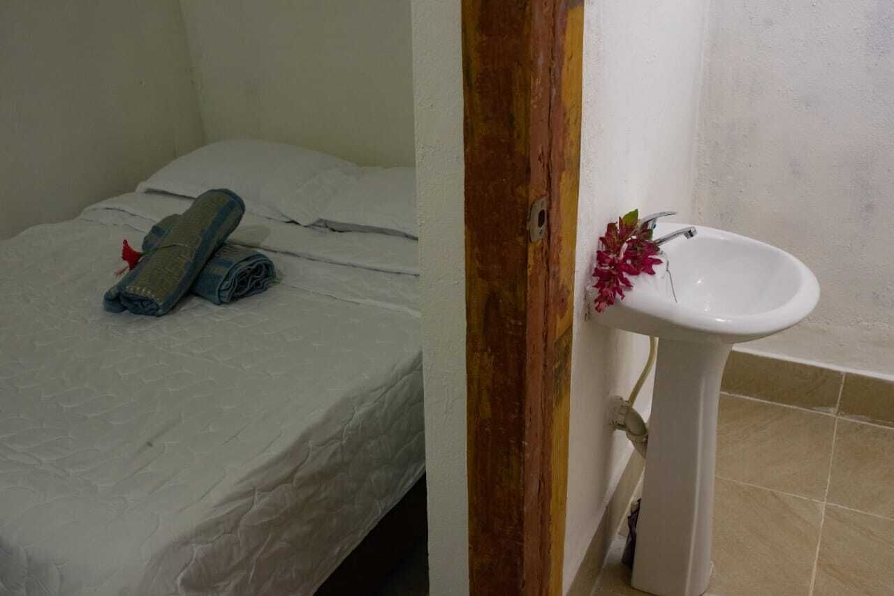 Una habitación pequeña con cama individual, colcha blanca, toalla enrollada y lavamanos blanco con un florero.