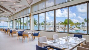 Un comedor amplio con ventanas grandes que dan a una piscina y palmeras. Las mesas están preparadas con manteles blancos y sillas azules.