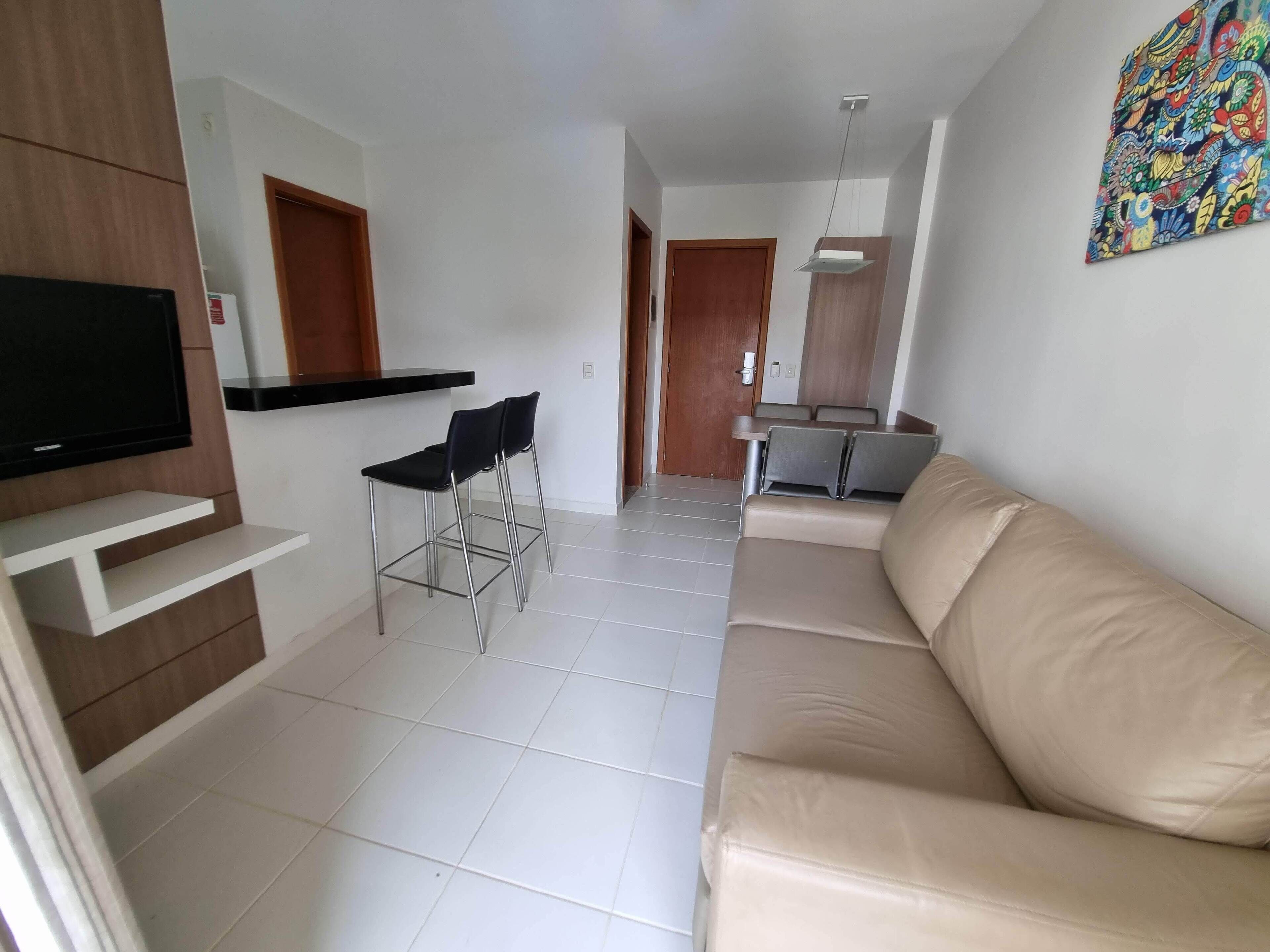 A modern living room with a beige sofa, a TV mounted on a wooden panel, and a dining area with chairs and a table.