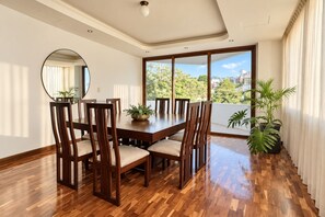 A dining area with a wooden table and chairs, large windows with a view of greenery and buildings, and a potted plant.