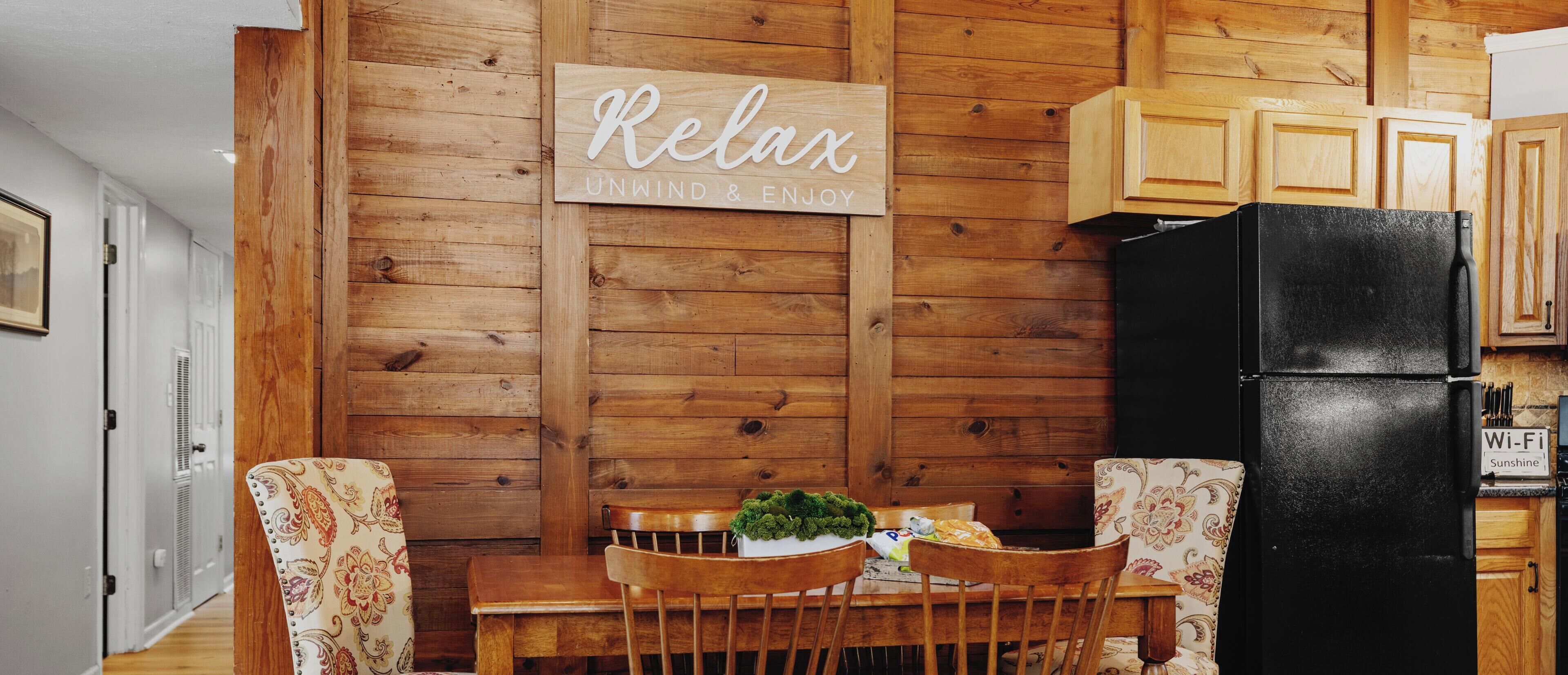 Une cuisine chaleureuse avec des murs en bois, un réfrigérateur noir et une table à manger avec des chaises. Une pancarte sur le mur porte l’inscription : « Détendez-vous, relaxez-vous et profitez de l’instant. »