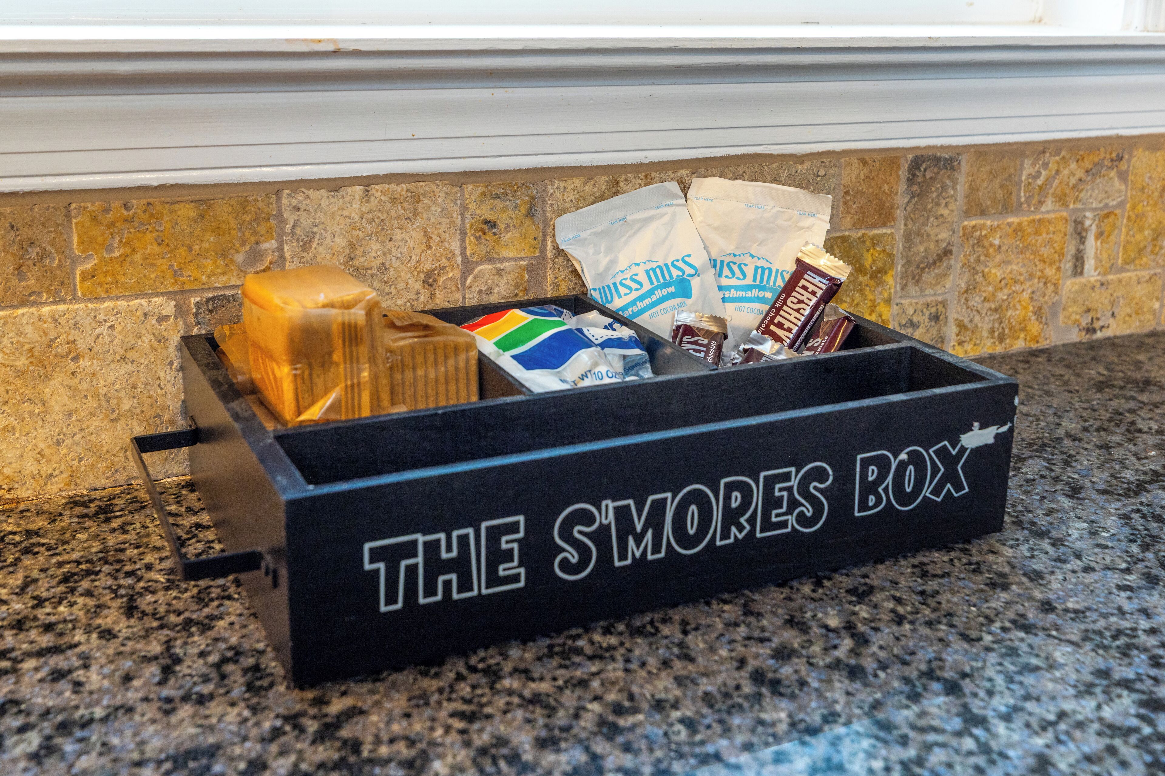 Une boîte noire portant l’inscription « THE SMORES BOX » est posée sur un comptoir en granit. Elle contient une guimauve, une tablette de chocolat et deux sachets de guimauves de la marque Miss Miss.