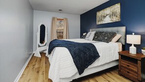 A bedroom with a bed, a nightstand, a lamp, a painting, and a window with curtains.