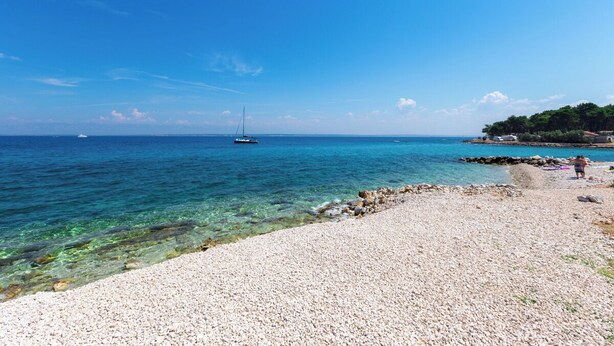A pebbled beach with clear turquoise water, a sailboat in the distance, and a rocky shoreline with a person standing. - Room-caravan Near the sea 4 (Preko)