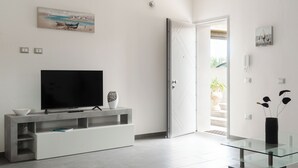 Een moderne woonkamer met een flatscreen televisie op een witte console, een glazen salontafel, en een deur die leidt naar een buitenruimte.
