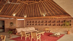 A room with a high, thatched ceiling, a wooden sofa, a dining table with chairs, and a brick wall with shelves. - Mariana Uno · Luxury · Exclusive Bay Oaxaca (Puerto Escondido)