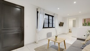 A modern living room with a dark wooden door, a window with white curtains, a small table with a vase, a grey sofa, and a cactus painting on the wall. - Bluestay 301 - Superb apartment in Paris (Paris 02 Bourse)
