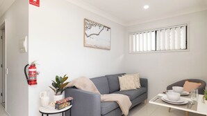 Una sala de estar moderna con un sofá gris, una mesita con un mantel blanco y una ventana con persianas. Un extintor de incendios está montado en la pared.