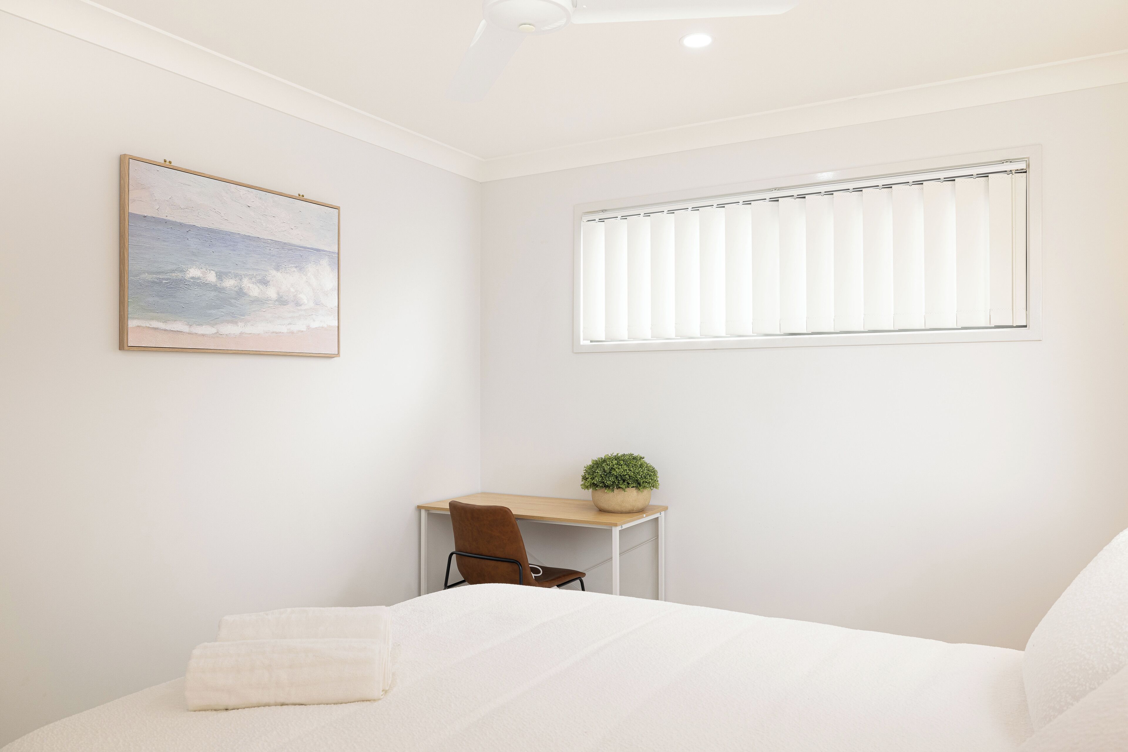 A bedroom with a bed, a desk with a chair, a window with blinds, and a painting on the wall.