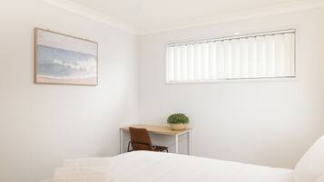 A bedroom with a bed, a desk with a chair, a window with blinds, and a painting on the wall.